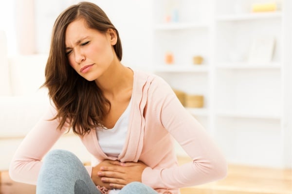 Woman sitting indoors, holding her stomach in discomfort, possibly experiencing abdominal pain or digestive issues.