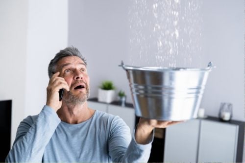 Concerned man holding a metal bucket to catch water leaking from the ceiling while talking on the phone, likely calling for repair assistance.