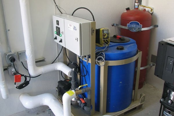 Hypochlorite skid-mounted chlorination system with blue chemical storage tank, metering pump, control panel, and connected PVC piping, installed indoors for water disinfection purposes.
