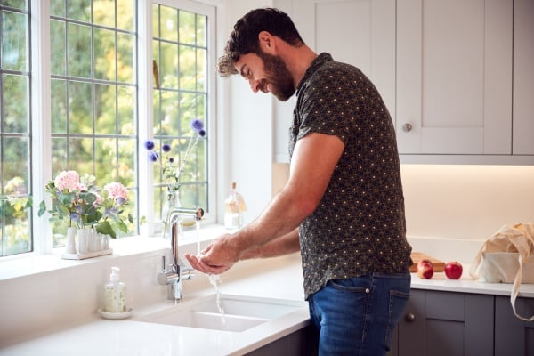 Bearded man washing hands under faucet in a bright modern kitchen.