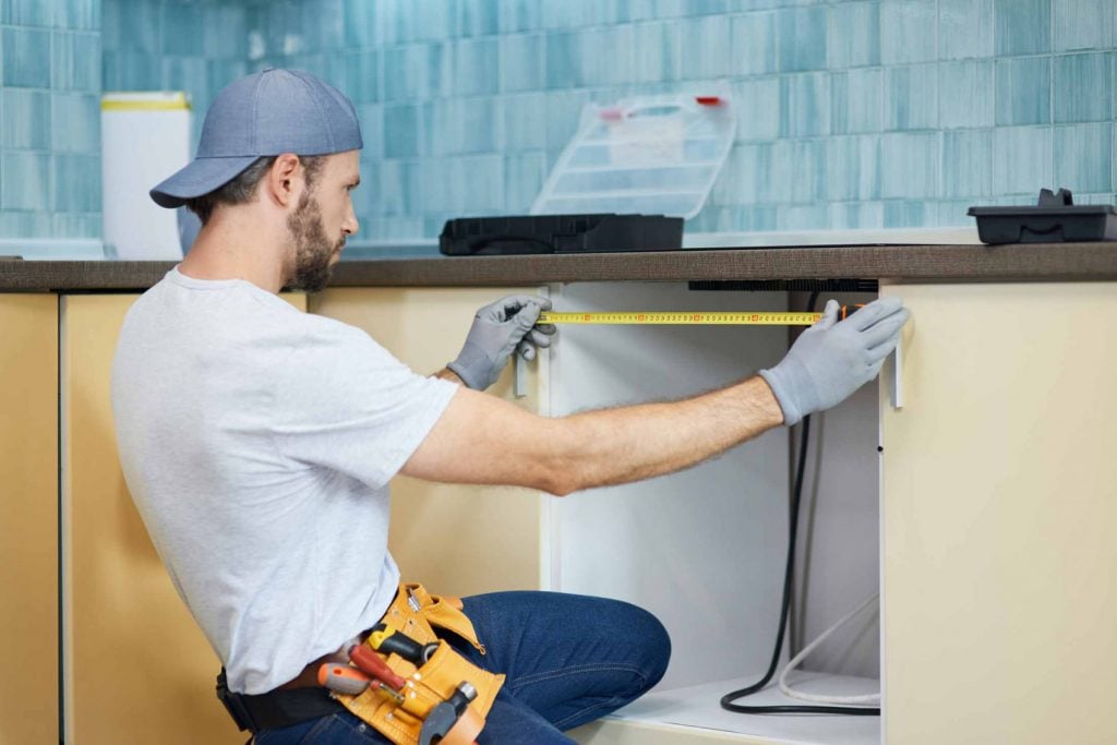 Plumber measuring under a kitchen sink cabinet to prepare for water filter installation.
