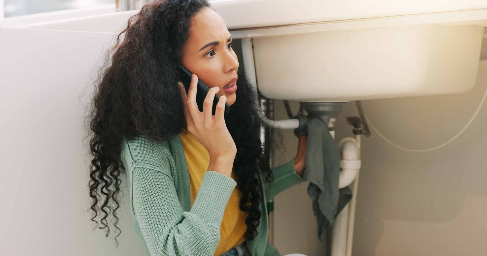 Worried homeowner calling a plumber while holding a towel under a leaking sink.