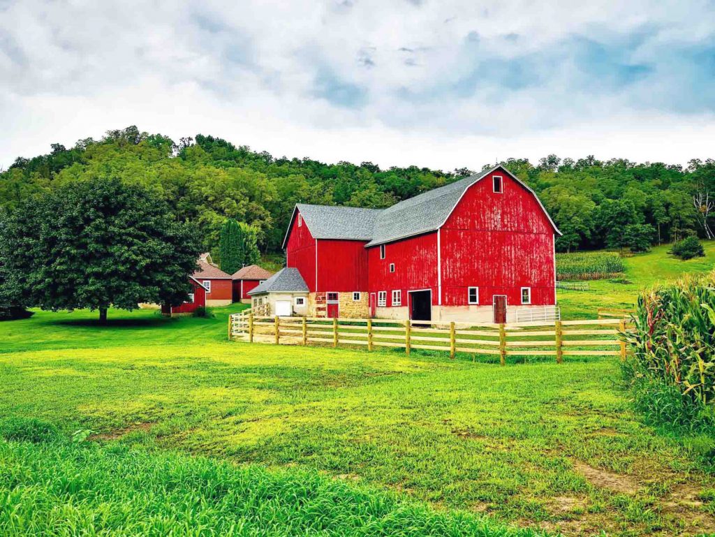 A classic red barn surrounded by green grass, trees, and farmland under a cloudy sky.