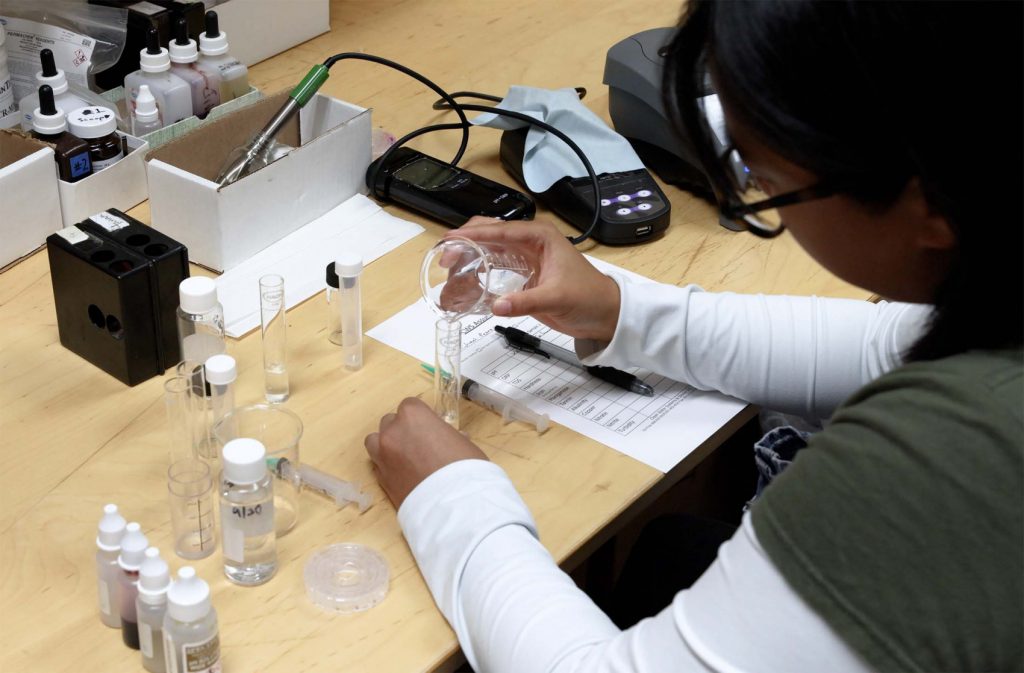 Technician in a lab testing water samples using glass beakers and instruments to measure water quality and contaminant levels.