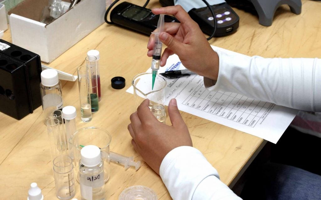 Technician using a syringe to test water samples in a lab with beakers, vials, and a printed results sheet on the table.