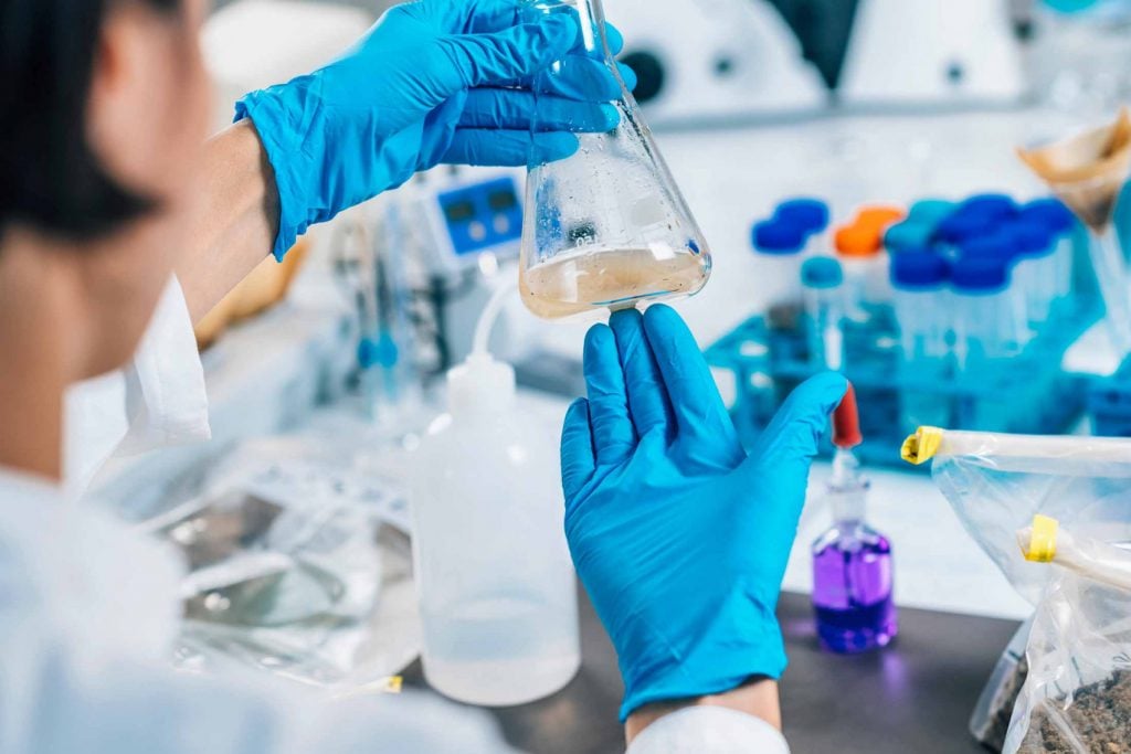 Lab technician wearing blue gloves analyzing a water sample in a glass flask to test for contaminants and clarity.