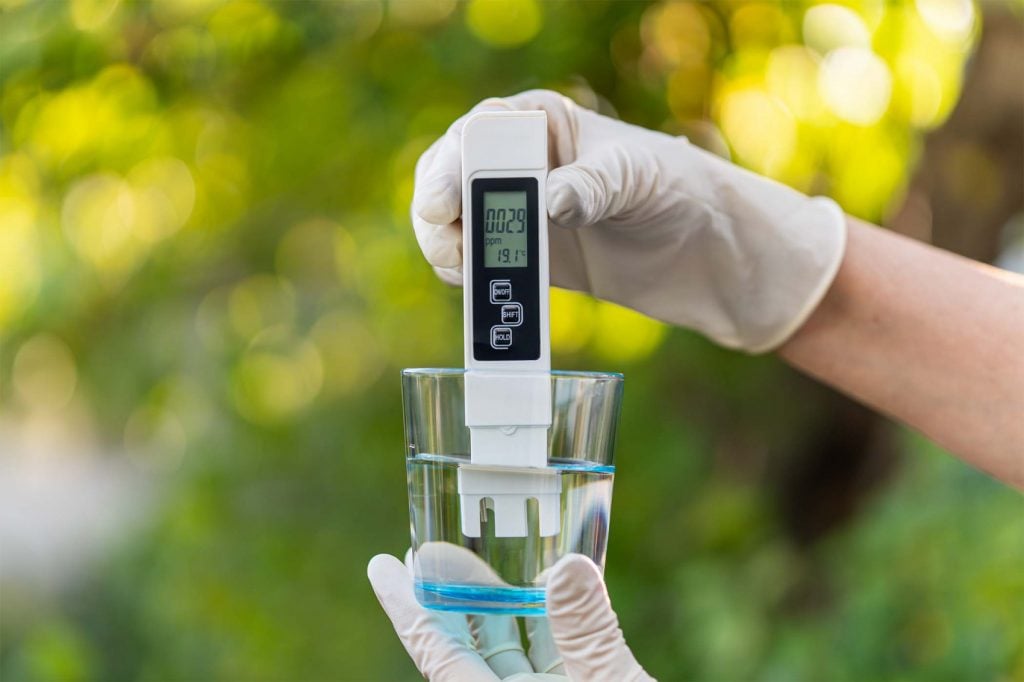 Gloved hand holding a digital TDS meter in a glass of water to test purity and total dissolved solids.