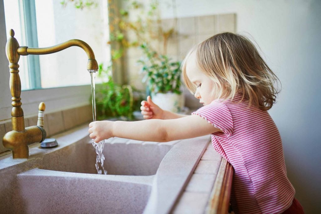 Young child standing at a kitchen sink with running tap water, symbolizing the importance of clean and safe household water.