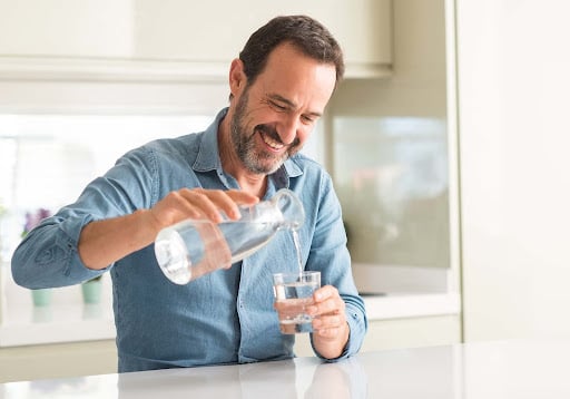 Smiling man in a blue shirt pouring water from a glass pitcher into a glass while standing in a bright kitchen, representing clean and refreshing drinking water.