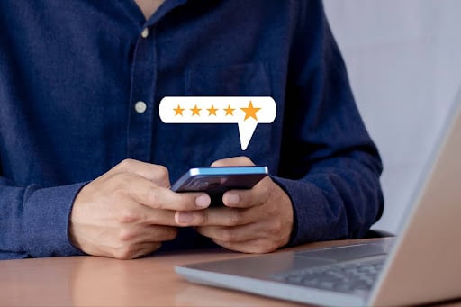 Person sitting at a desk using a smartphone with a five-star rating graphic appearing above the device, representing customer feedback or online reviews.