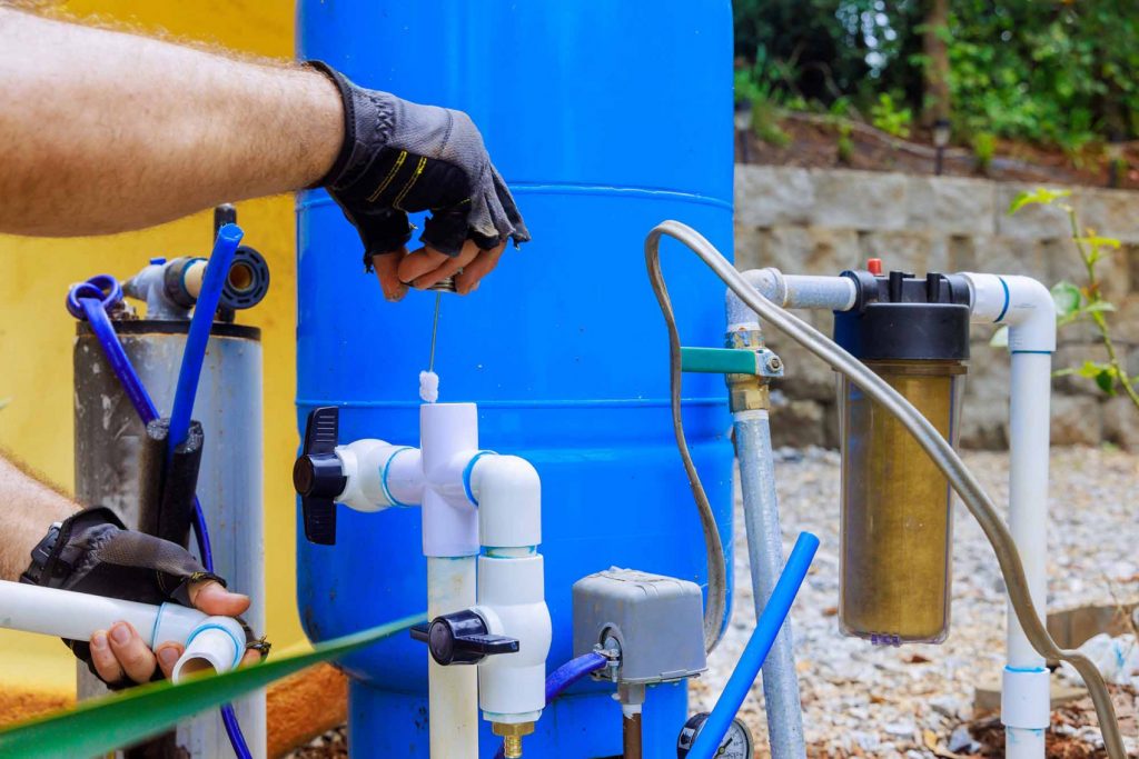 Technician performing maintenance on a home well water system, inspecting PVC connections and filtration components near a blue pressure tank.