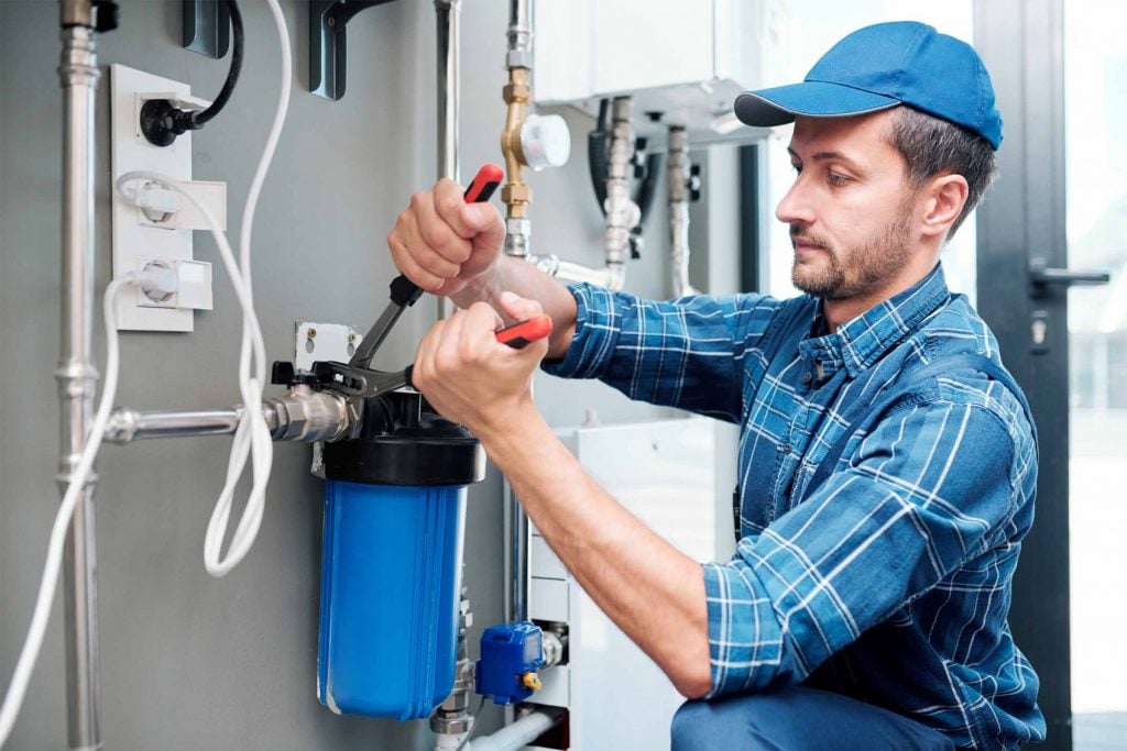 Plumber using a wrench to install a blue whole-house water filter canister for a residential well water system.