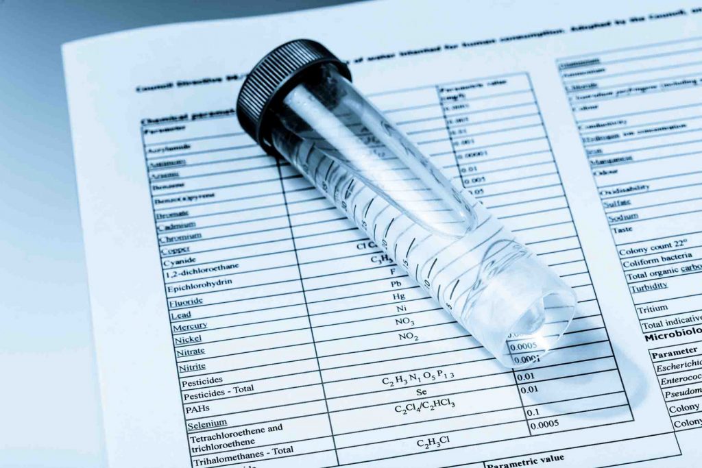 Clear water sample vial placed on a printed laboratory water quality report showing contaminant levels and test results for home drinking water.