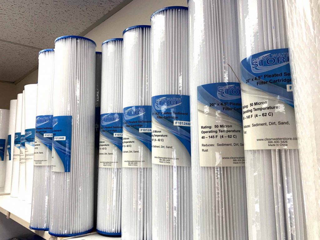 Rows of 20" x 4.5" pleated sediment filter cartridges from Clean Water Store displayed on a shelf. These filters remove sediment, dirt, sand, and rust from well or city water systems to protect plumbing and appliances.
